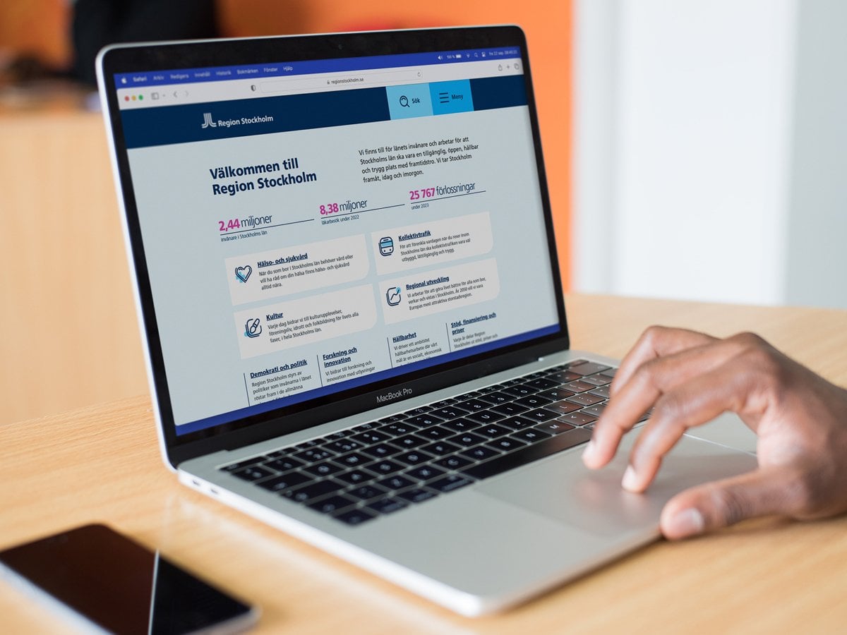 Foto på en laptop som står på ett bord. En person som inte syns i bild har sin hand på musplattan. På skärmen är Region Stockholms testversion av sin nya webbsida öppen. Till vänster om datorn ligger en mobiltelefon på bordet.