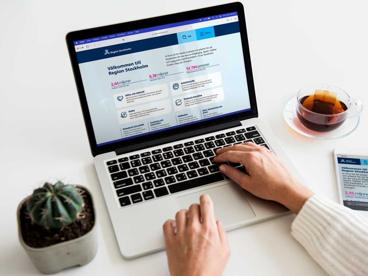 Foto på en person som använder en dator och har en mobiltelefon bredvid datorn. På datorn och mobiltelefon är Region Stockholms webbsida öppen