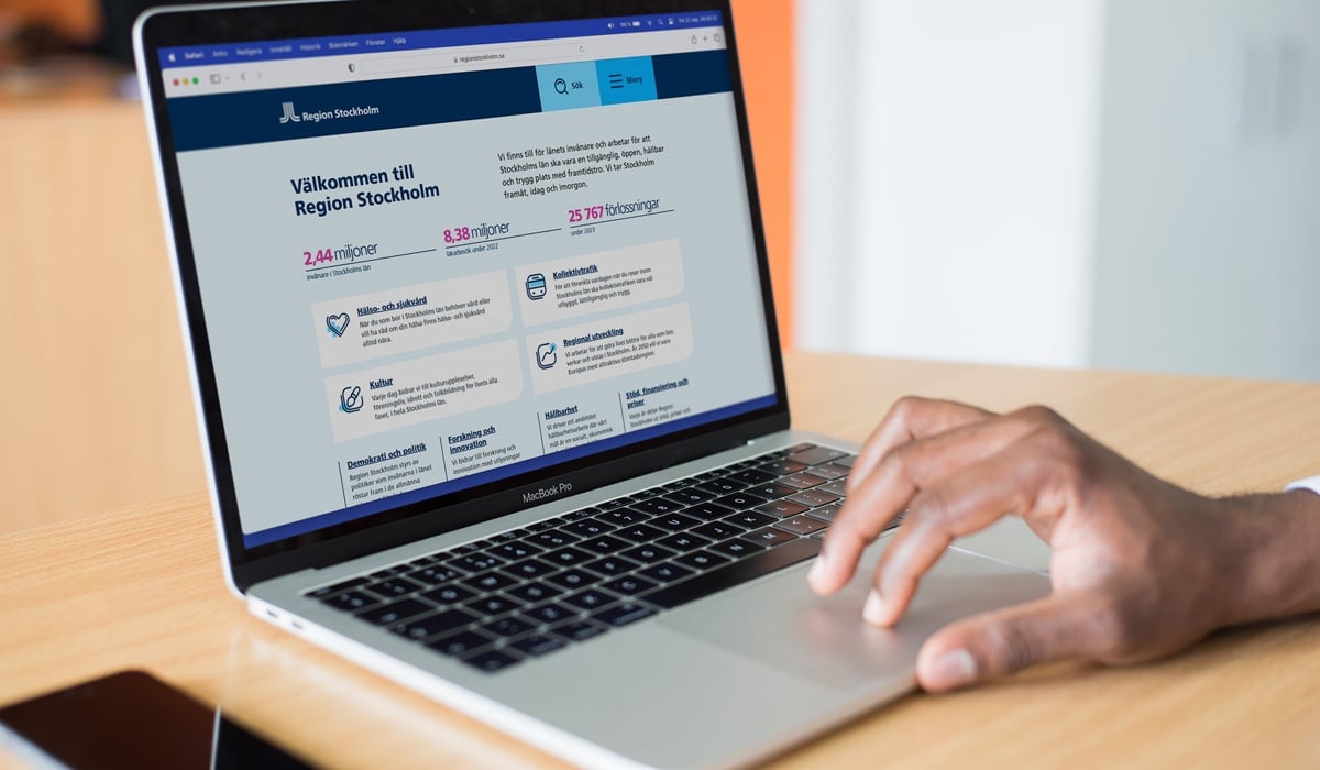 Foto på en laptop som står på ett bord. En person som inte syns i bild har sin hand på musplattan. På skärmen är Region Stockholms testversion av sin nya webbsida öppen. Till vänster om datorn ligger en mobiltelefon på bordet.