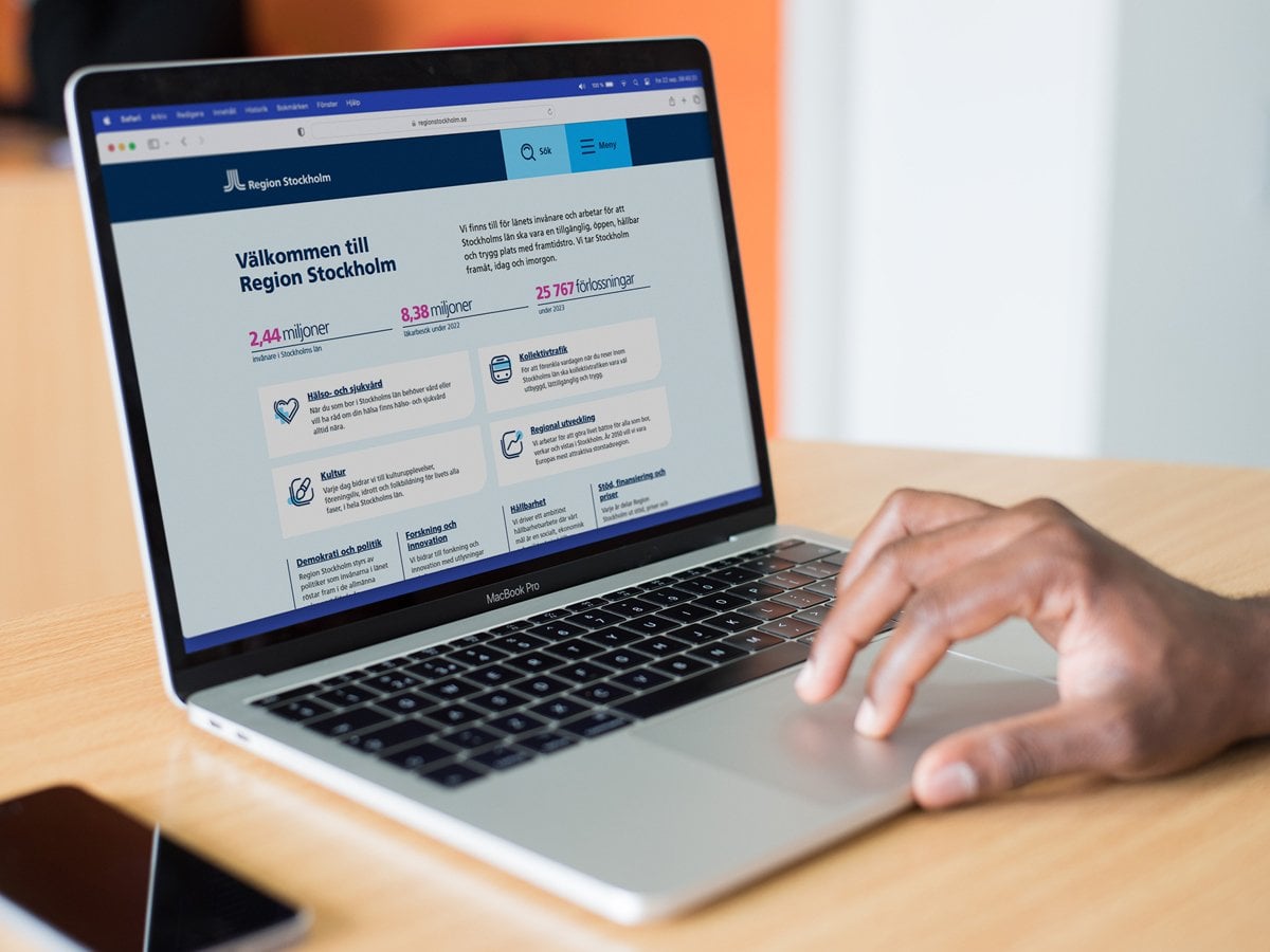 Foto på en laptop som står på ett bord. En person som inte syns i bild har sin hand på musplattan. På skärmen är Region Stockholms testversion av sin nya webbsida öppen. Till vänster om datorn ligger en mobiltelefon på bordet.
